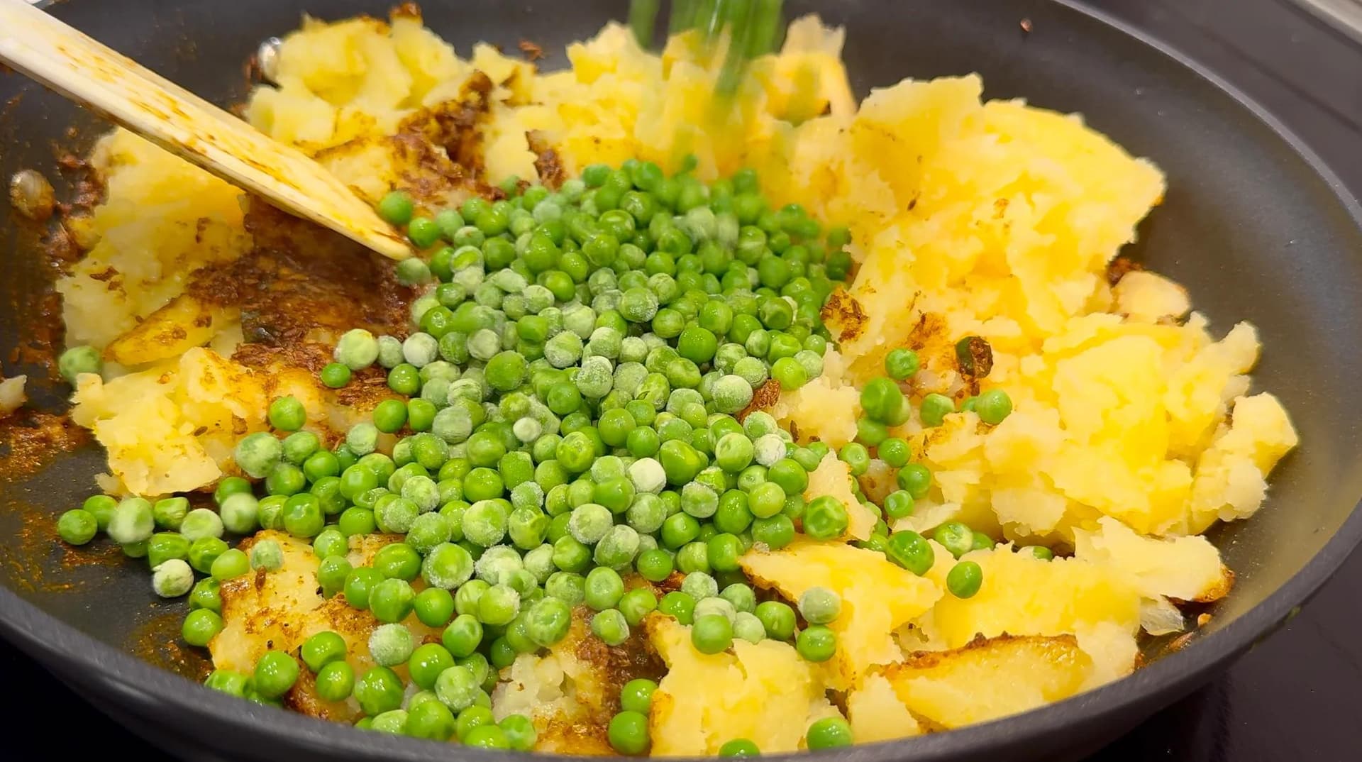 Diced potatoes and peas being stirred together in a dark pan with spices during samosa filling preparation.