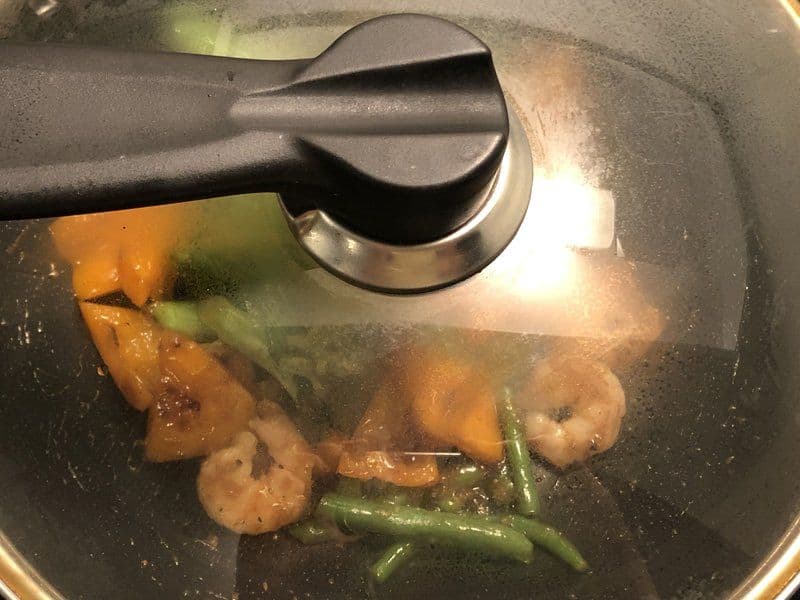 Stir-frying shrimp and green beans in a dark pan with a lid partially closed during cooking.