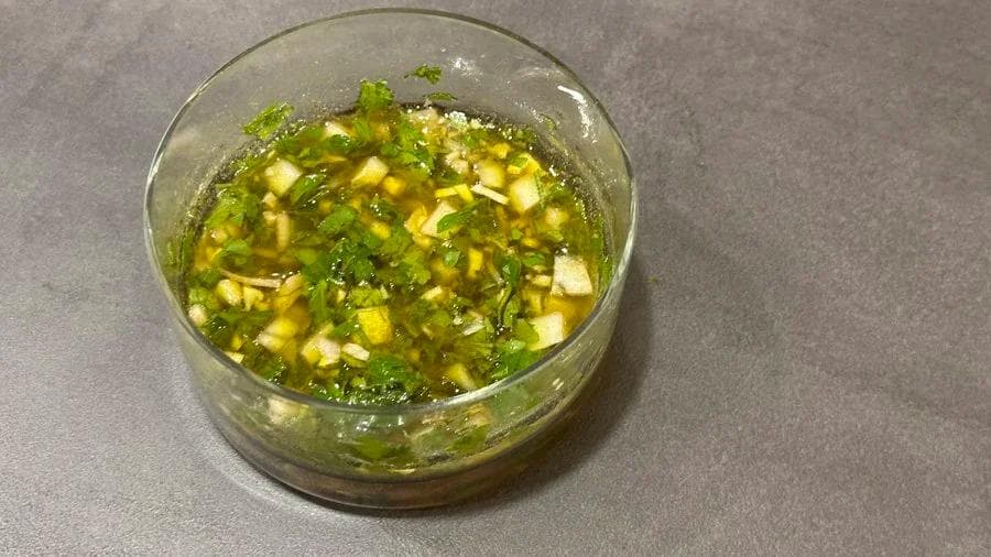 Green chimichurri sauce in a clear glass bowl with herbs, garlic, and oil mixed together