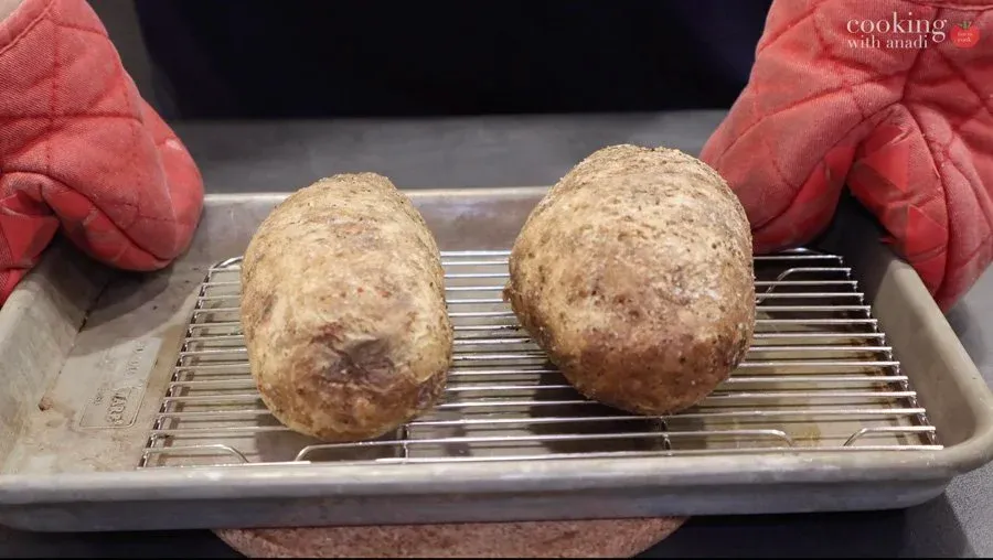 Two baked potatoes with crispy golden-brown skins on a cooling rack, ready to be checked for doneness.