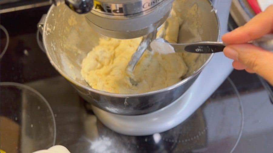 Stand mixer bowl with thick yellow cookie batter being scraped with a spatula during mixing.