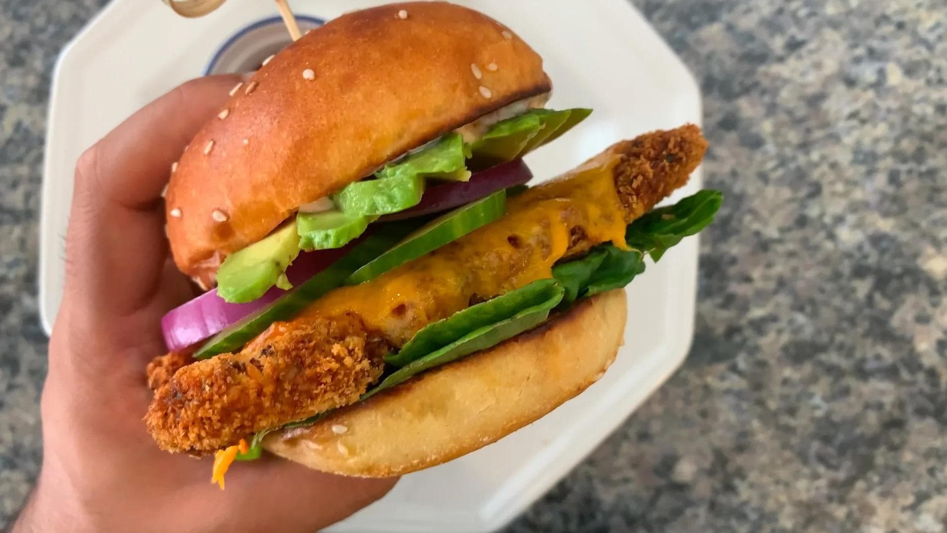 Hand holding a crispy panko chicken burger with lettuce, cucumber, onion, and avocado on a seeded bun.