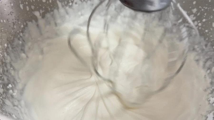 Electric mixer whipping heavy cream, powdered sugar, and vanilla extract to soft peaks in a bowl.