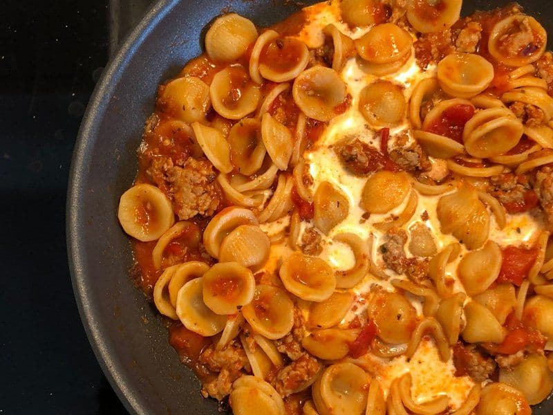 Orecchiette pasta with spicy Italian sausage and tomato sauce cooking in a skillet, topped with cream.