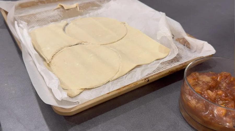 Puff pastry circles with pencil-drawn filling guides on parchment paper next to apple pie filling in a jar.