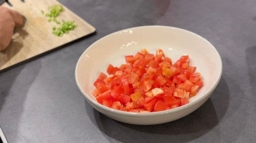 Diced red tomatoes in a white bowl on a gray countertop with chopped green onions on a cutting board nearby.