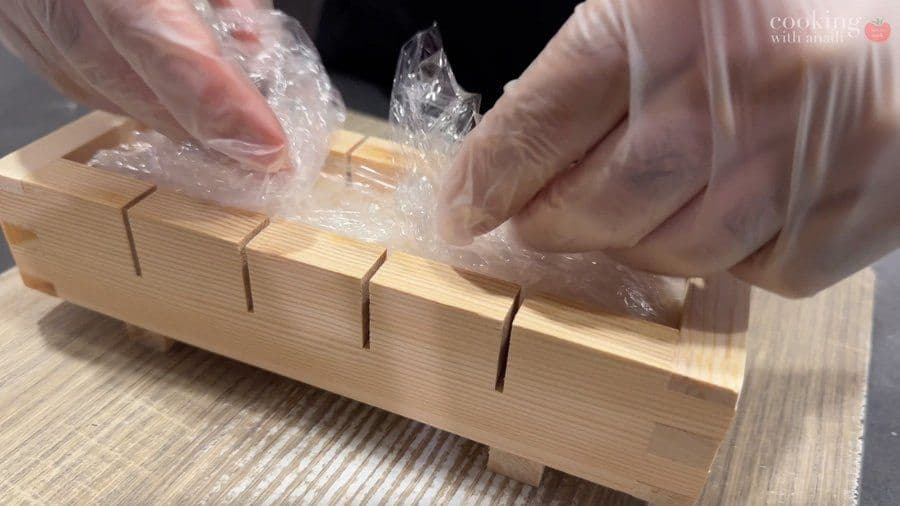 Hands in plastic gloves cutting through pressed sushi block with pre-marked guides using a sharp knife.