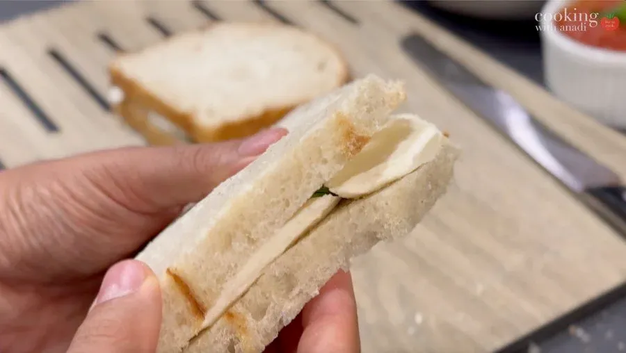Hand holding a bread-crusted mozzarella sandwich with melted cheese visible between white bread slices.