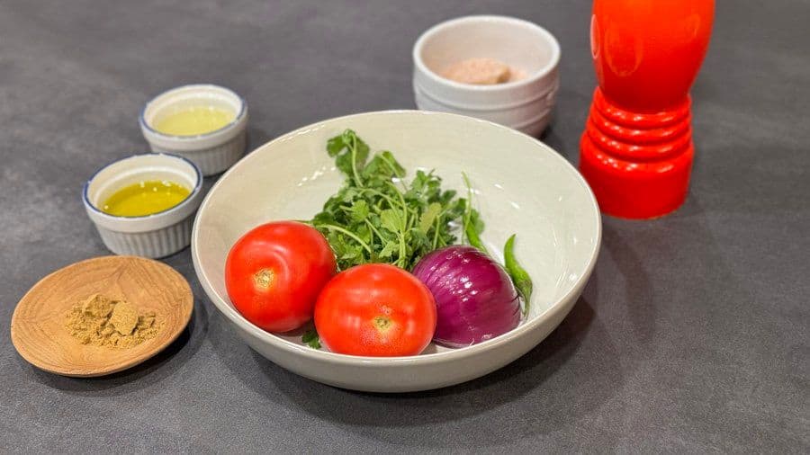 Ingredients for pico de gallo arranged in a white bowl: fresh tomatoes, red onion, cilantro, with oils and spices on the side.