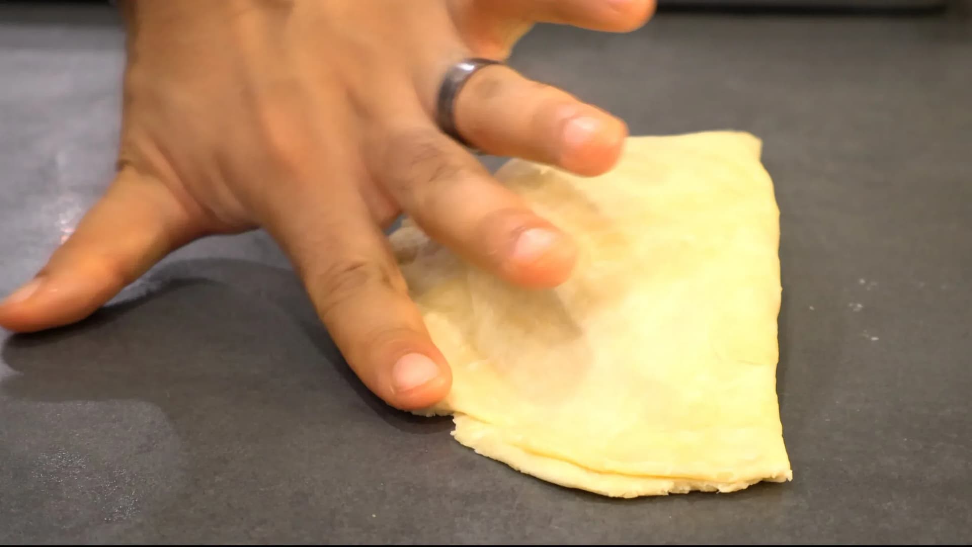Hand folding yellow dough over filling to create a handheld meat pie on dark surface.