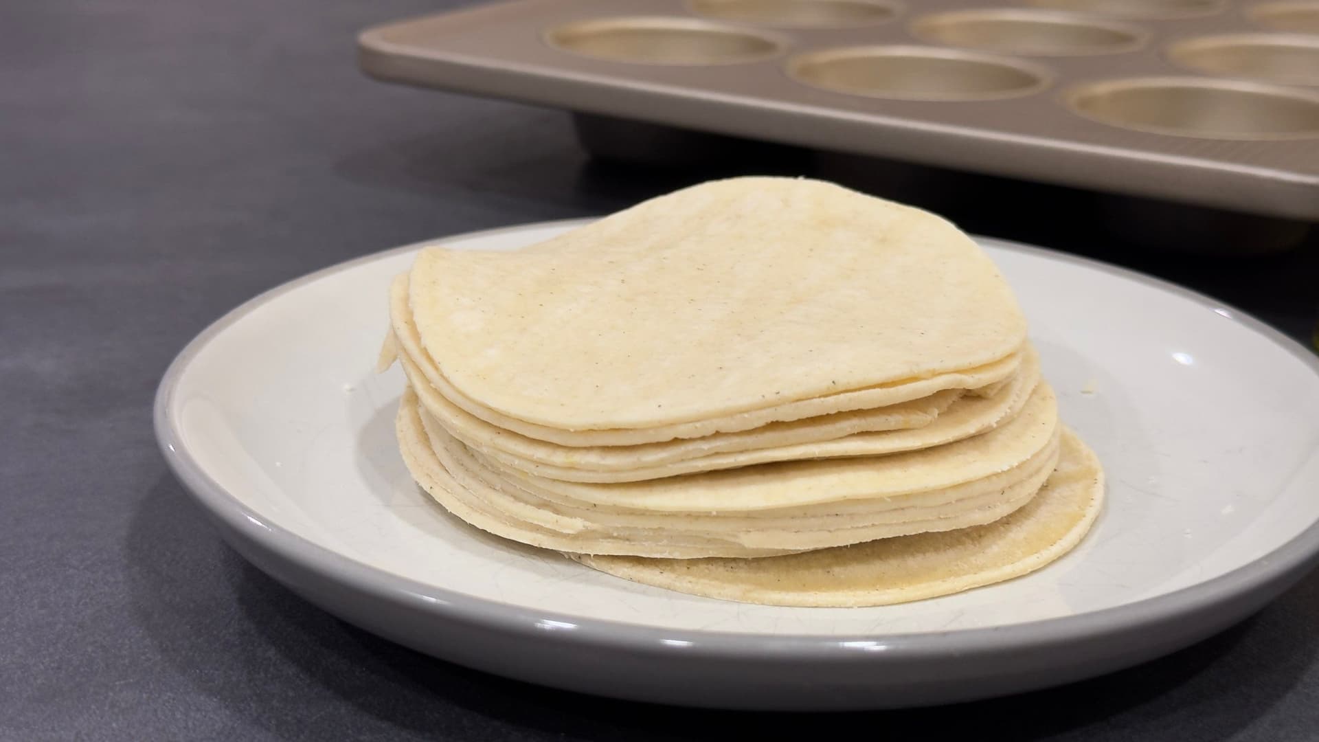 Stack of thin corn tortillas on a white plate with a muffin tin visible in the background