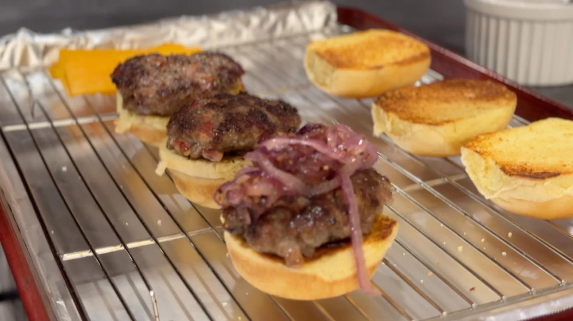 Bacon cheeseburger sliders on toasted buns with caramelized onions, served on a wire cooling rack.
