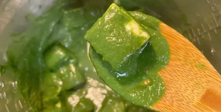 Green bell pepper being lifted on wooden spoon from simmering palak paneer curry in pot.