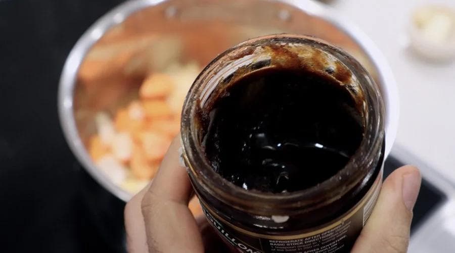 A hand holding a jar of dark Better Than Bouillon broth concentrate over a pot of carrot and ginger soup ingredients.