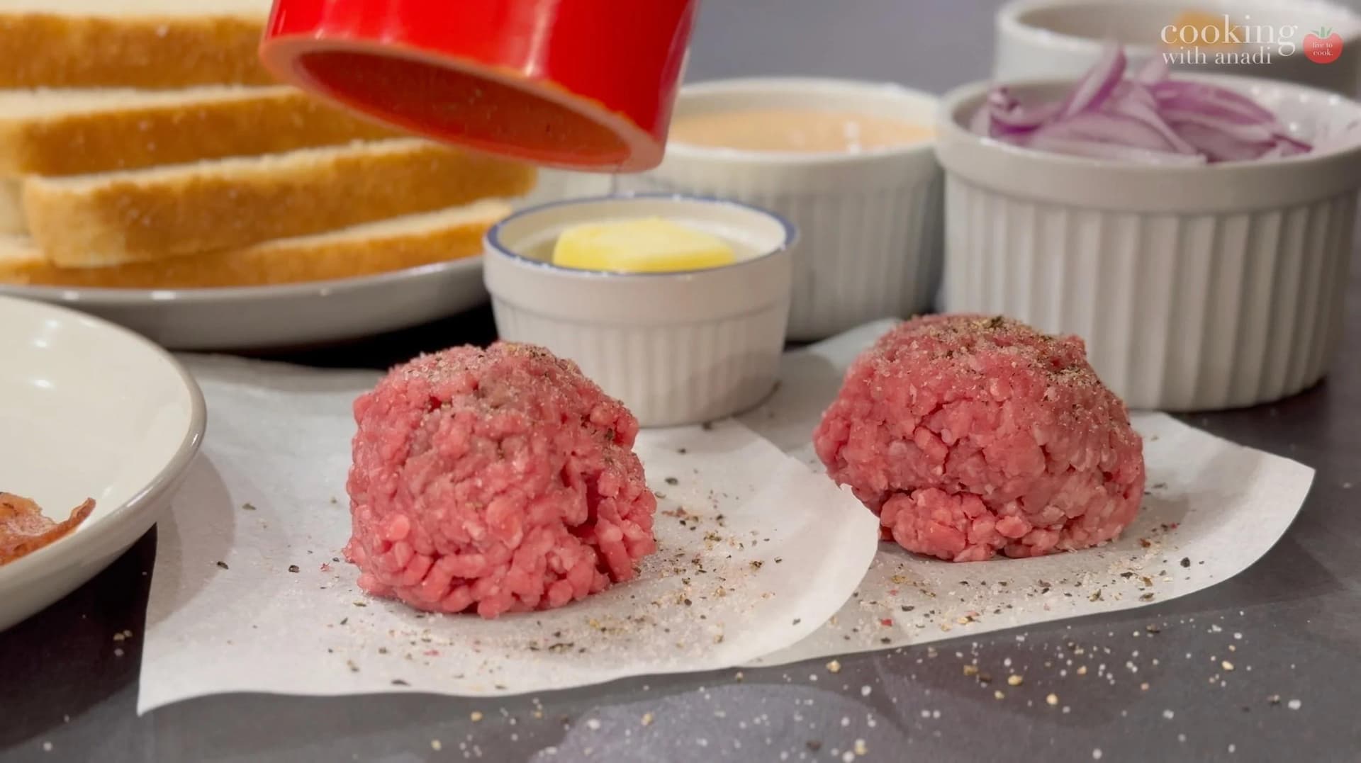 Two pink ground beef patties seasoned with salt and pepper on parchment paper, surrounded by burger ingredients including toasted bread and butter.