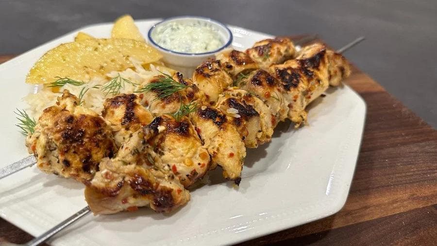 Grilled Greek souvlaki chicken skewers with charred edges, served with tortilla chips and tzatziki sauce on a white plate.