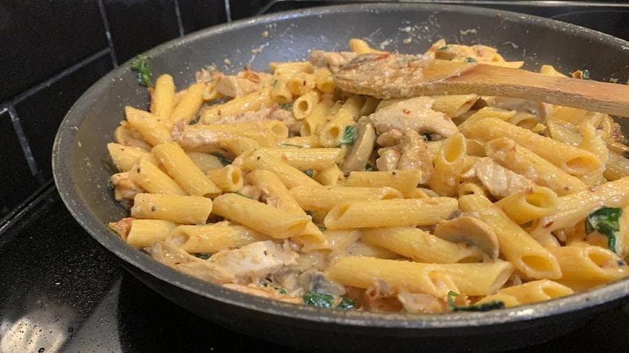 Spicy creamy chicken penne pasta in a skillet with wooden spoon, garnished with fresh herbs.