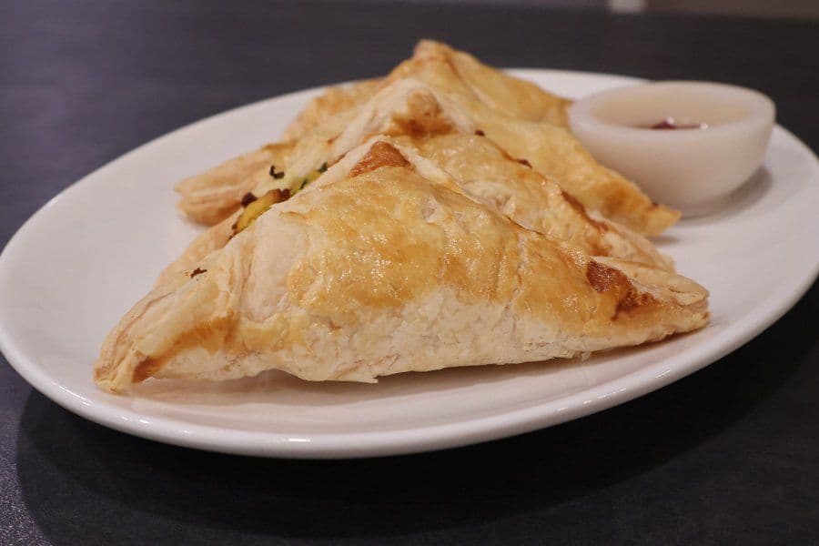 Two golden-brown aloo puff pastry patties on a white plate with a small bowl of ketchup.