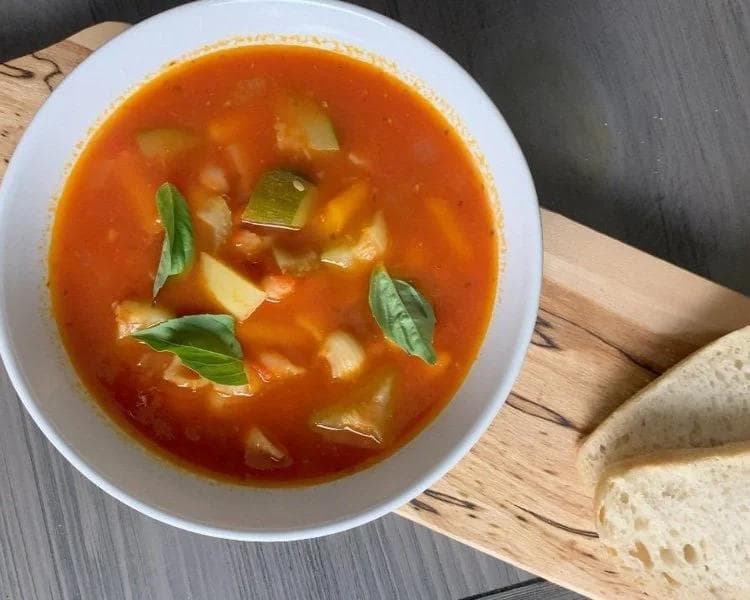 Bowl of vegan minestrone soup with red broth, vegetables, and fresh basil garnish, served with bread.