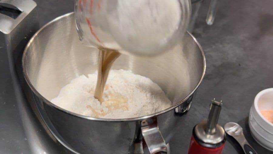 Pouring yeast mixture into flour in a stand mixer bowl during pizza dough preparation.