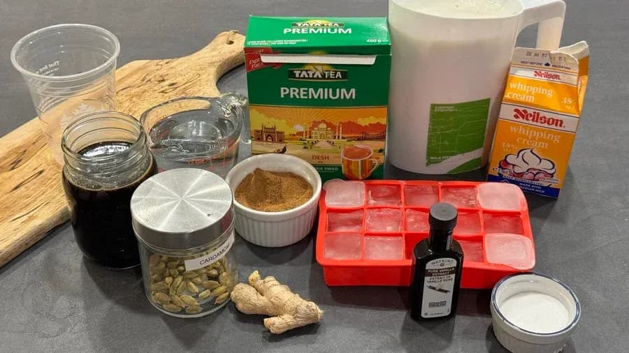 Ingredients for iced gingerbread chai latte laid out on gray surface, including tea box, spices, milk, cream, peanuts, and storage containers.