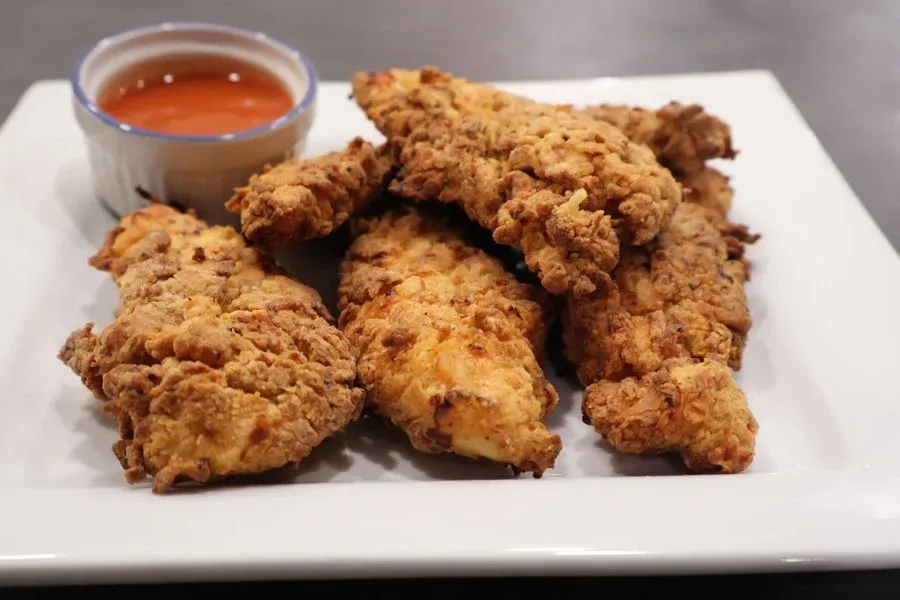 Crispy golden fried chicken tenders piled on a white plate with a small ramekin of red dipping sauce.