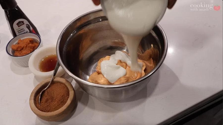 Pouring sour cream into a bowl of pumpkin cream cheese filling ingredients with cinnamon and other seasonings visible.
