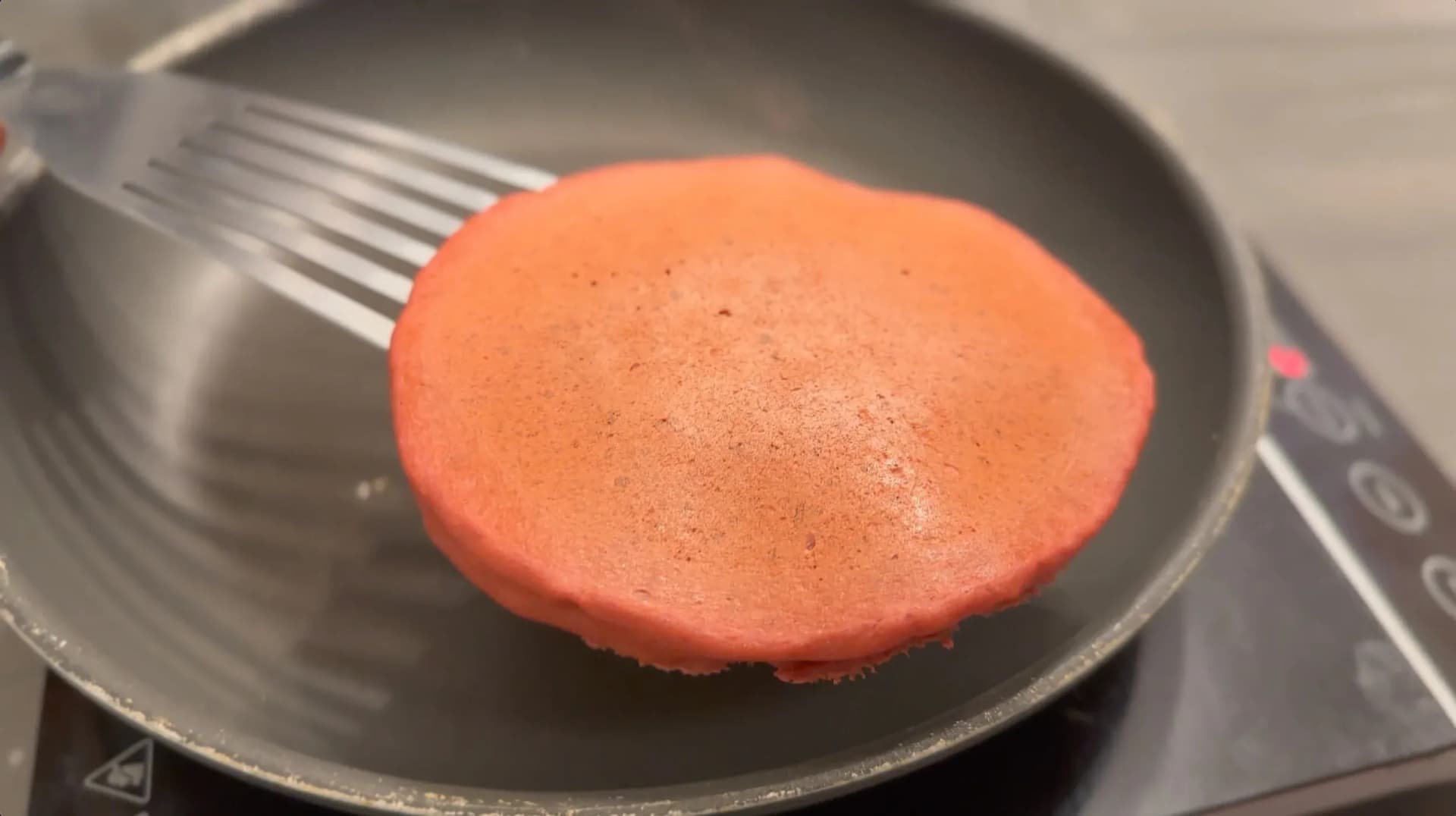 A red velvet pancake cooking in a dark skillet with a spatula, showing bubbles forming on the surface.