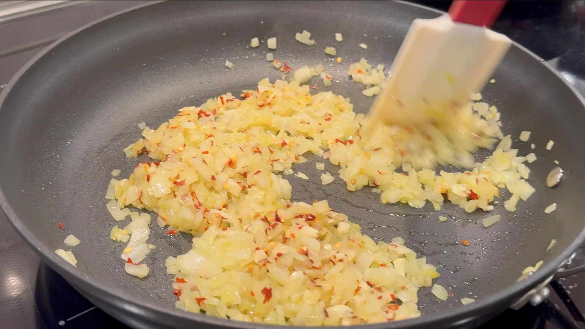 Diced onions, garlic, and red chilli flakes sautéing in a non-stick pan, being stirred with a white spatula.