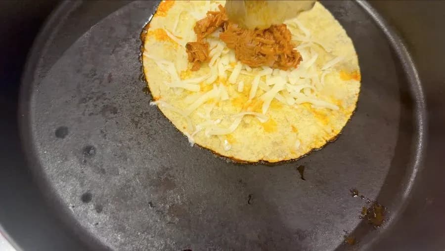 A birria taco searing in a non-stick pan with melted cheese and shredded meat on a dipped tortilla.