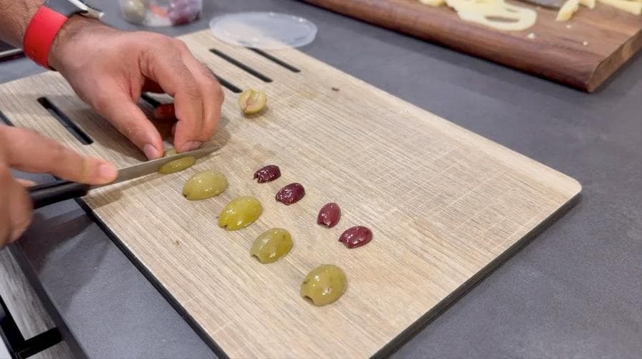 Halved olives and olive strips arranged on a wooden cutting board for making olive spider pizza toppings.
