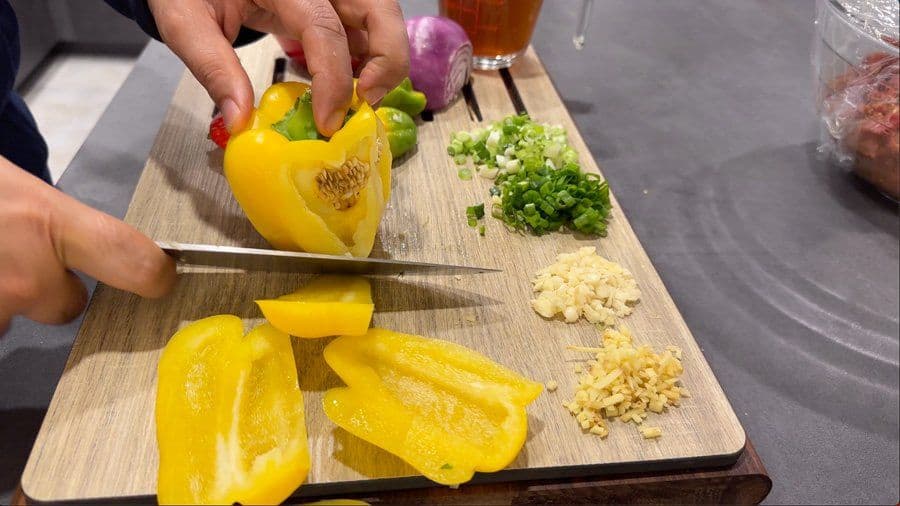 Hands chopping yellow bell peppers on a cutting board with minced garlic, ginger, and green onions nearby.