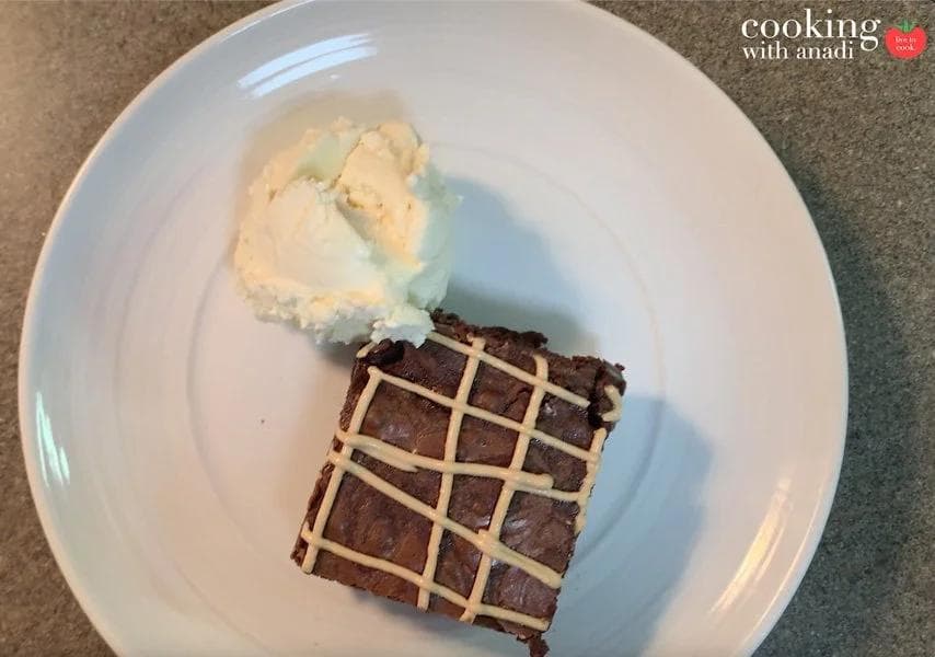 Espresso brownie square with white drizzle topped with a scoop of peppermint mocha cream on white plate.