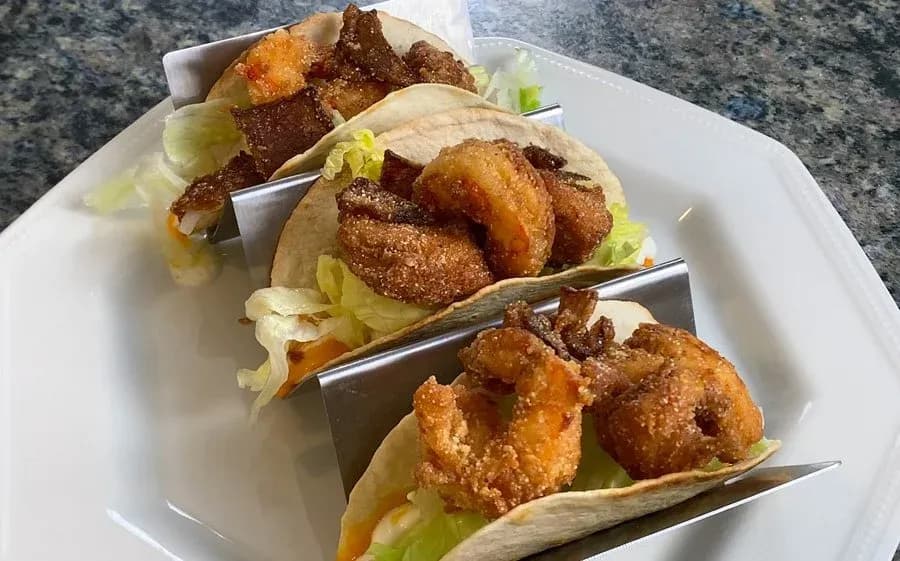 Three crispy chicken and shrimp tacos in small tortilla shells with shredded lettuce, served on a white plate.