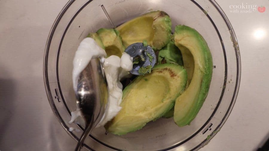 Sliced avocados and sour cream in a food processor bowl ready to blend into creamy sauce.