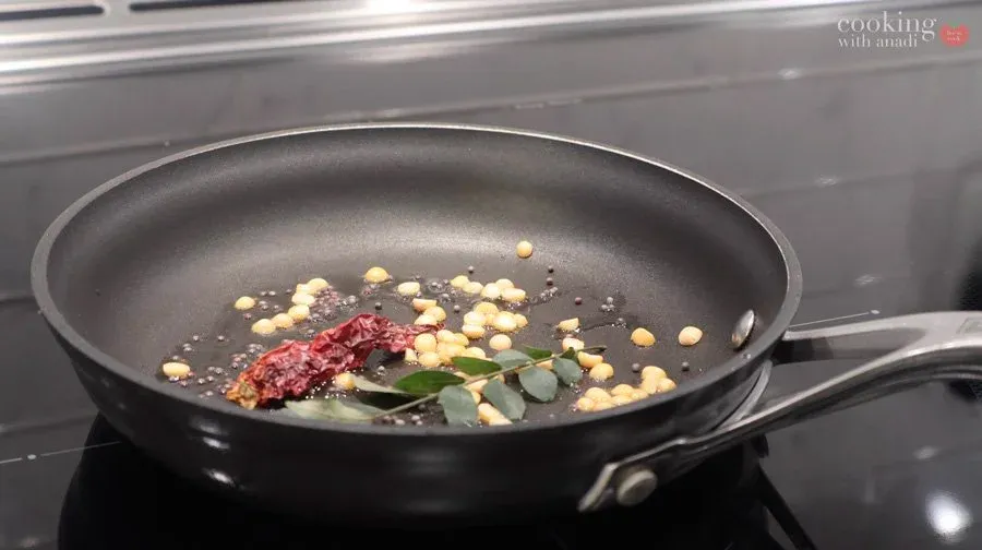 Tempering spices in a black pan: mustard seeds, chana dal, dried red chilies, curry leaves, and hing with oil.